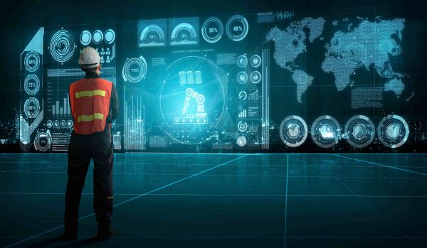 **Alternativtext:** Eine Person mit Schutzhelm und Warnweste steht vor einer großen digitalen Benutzeroberfläche mit Diagrammen, Weltkarte, Kennzahlen und einem Roboterarm-Symbol. Das Motiv visualisiert Industrie 4.0, digitale Transformation, KI und datenbasierte Steuerung in der Produktion.