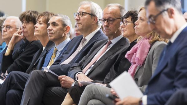 Friedrich Merz sitzt mit weiteren Teilnehmenden in der ersten Reihe einer Veranstaltung und blickt gedankenversunken nach unten.